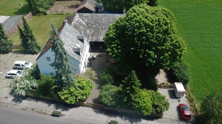 Un grand terrain vert avec une maison blanche et un grand arbre. Autour de la maison, on peut voir quelques voitures et beaucoup de verdure.