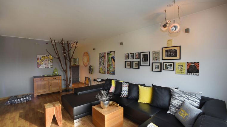 Ein modernes Wohnzimmer mit einem großen schwarzen Sofa und Kunstwerken an den Wänden. Es gibt einen Holzschrank und einen Tisch aus Holz sowie eine stilvolle Beleuchtung.