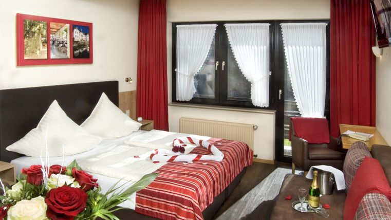 Ein gemütliches Hotelzimmer mit einem großen Bett und roten Vorhängen. Auf dem Nachtisch liegen Blumen und es gibt eine Flasche Sekt bereit.