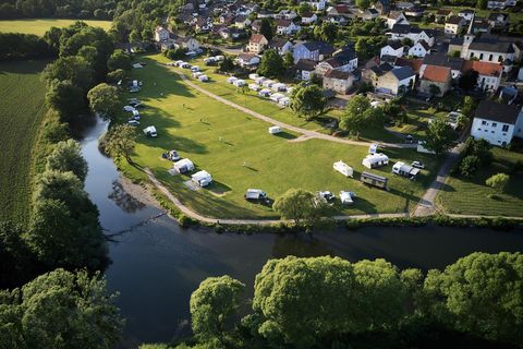Ein Campingplatz am Fluss mit vielen Wohnmobilen und Zelten. Im Hintergrund sind Häuser und eine grüne Landschaft zu sehen.