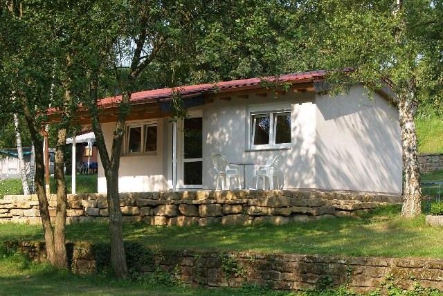 Ein kleines, gemütliches Ferienhaus mit rotem Dach, umgeben von Bäumen und grüner Wiese. Es gibt zwei Stühle vor dem Eingang, ideal für Entspannung im Freien.