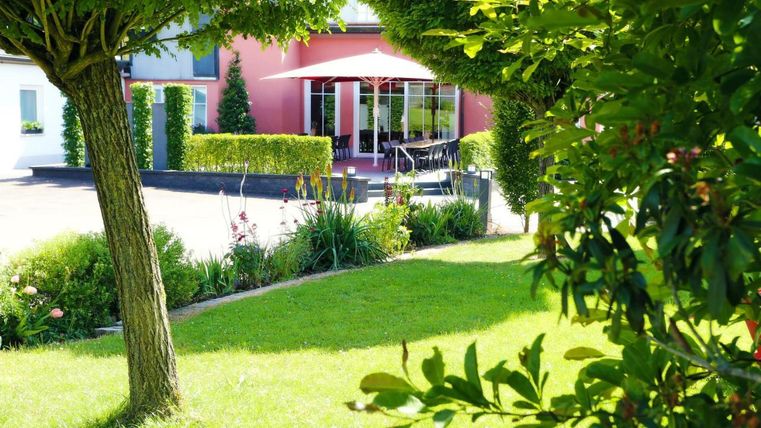 Ein gepflegter Garten mit grünen Hecken und Bäumen. Im Hintergrund ist ein Gebäude mit einem Terrasse und Sonnenschirm sichtbar.