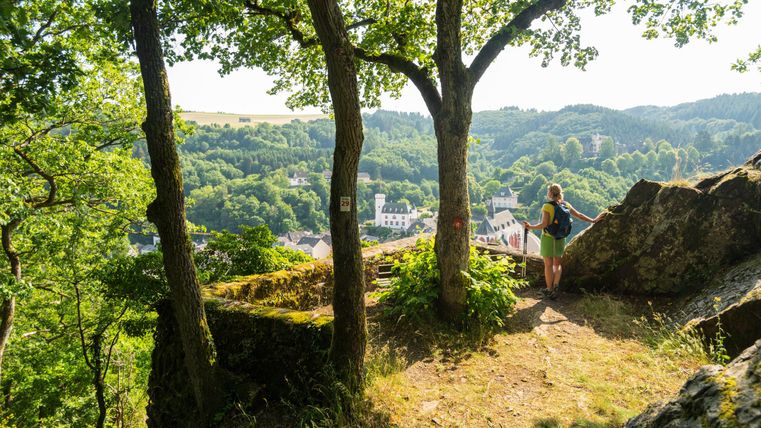 Eine Wandererin steht auf einem Felsen und blickt auf ein malerisches Tal mit grünen Bäumen und kleinen Häusern. Die Sonne scheint und der Himmel ist klar.