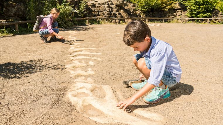 Zwei Kinder spielen in einem Sandfeld. Ein Junge zeichnet etwas in den Sand, während ein Mädchen im Hintergrund zuschaut.