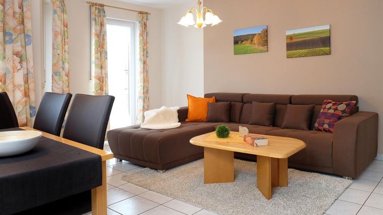 Ein helles Wohnzimmer mit einem gemütlichen Sofa und einem Holztisch. An den Wänden hängen Bilder und die Fenster sind mit bunten Vorhängen dekoriert.