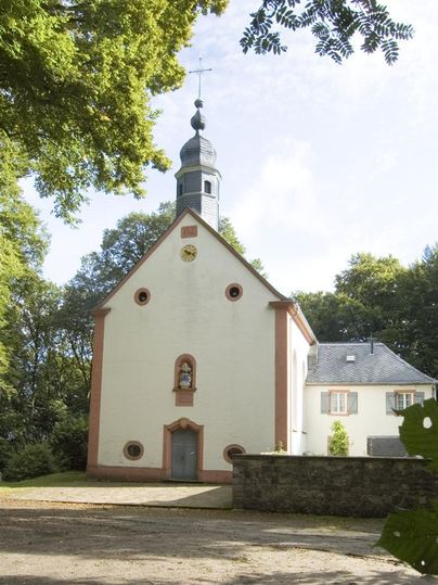 Eine kleine, charmante Kirche mit einem Turm, umgeben von Bäumen. Die Fassade ist weiß mit roten Akzenten und hat einladende Doppel-Fenster.