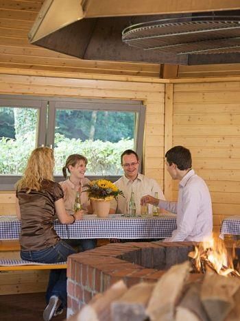 Eine Gruppe von vier Personen sitzt in einer gemütlichen Hütte und genießt Zeit miteinander. Auf dem Tisch steht ein Blumenstrauß und im Hintergrund brennt ein Kamin.