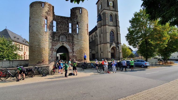 Gruppe von Radfahrern vor einem historischen Tor und einer Kirche.