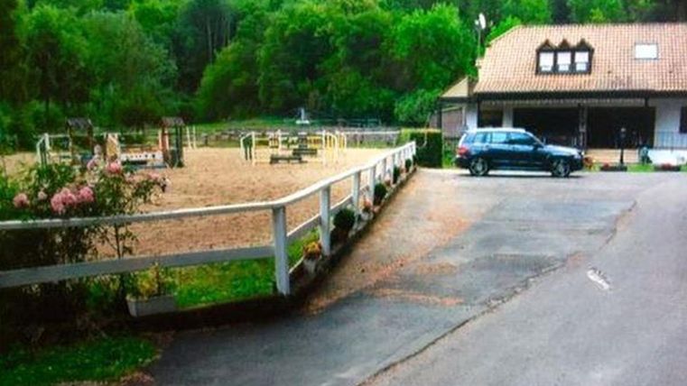 Ein Reitplatz mit Hindernissen und ein angrenzendes Gebäude sind sichtbar. Die Umgebung ist grün und von Bäumen umgeben.