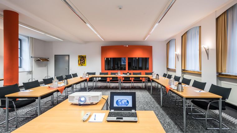 Ein moderner Konferenzraum mit Tischen und Stühlen in U-Form. An der Wand befindet sich ein Beamer und ein Bildschirm, und der Raum hat große Fenster mit Tageslicht.