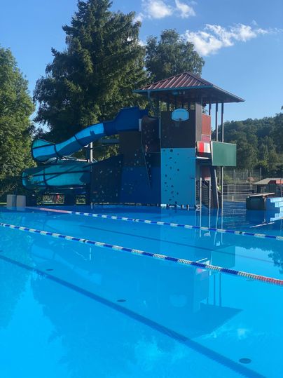 Ein Schwimmbad mit einem blauen Wasserrutsche und einem Spielplatzbereich. Die Wasseroberfläche ist klar und spiegelt den blauen Himmel wider.