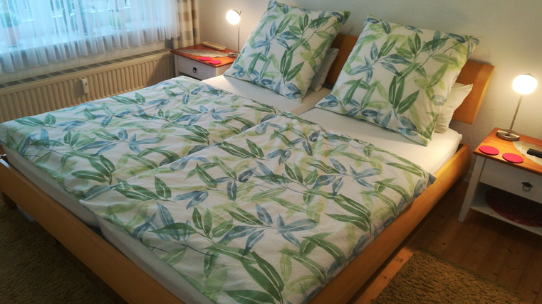 Ein gemütliches Schlafzimmer mit einem Doppelbett und dekorativer Bettwäsche in grünem Blattdesign. Der Raum ist hell erleuchtet und wirkt einladend.