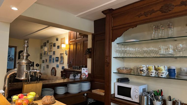 Une cuisine chaleureuse avec des étagères en bois, affichant de la vaisselle et des verres. Sur le plan de travail, on trouve des fruits frais et du pain, et en arrière-plan, des images sont accrochées au mur.