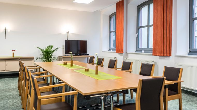 Ein moderner Konferenzraum mit einem langen Tisch, Holzstühlen und großen Fenstern. An der Wand befindet sich ein Fernseher und einige Pflanzen sorgen für eine angenehme Atmosphäre.