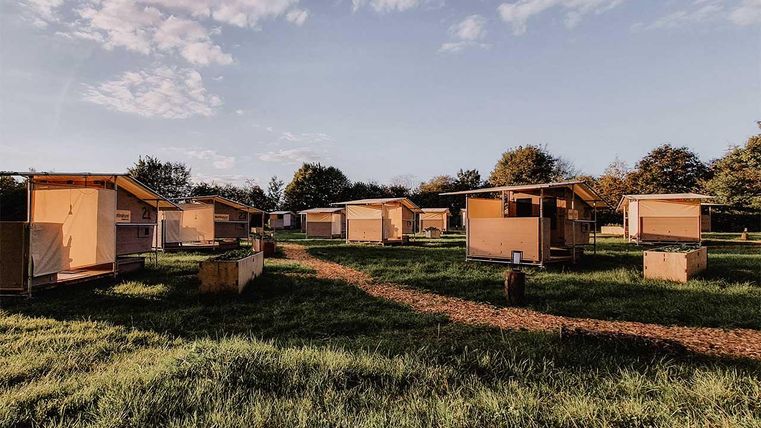 Eine Gruppe von Holzhütten steht in einem grünen Feld. Der Himmel ist leicht bewölkt und der Boden hat einen Weg aus Kies.