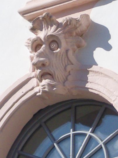 Ein dekorativer Fassadenelement in Form eines Löwenkopfes mit ausgeprägten Zügen. Das Relief ist in einem hellen Farbton gehalten und befindet sich über einem Fenster.