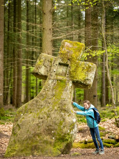 Ein Mädchen lehnt sich an das Fraubillenkreuz, ein moosbedecktes Steinkreuz im Wald.