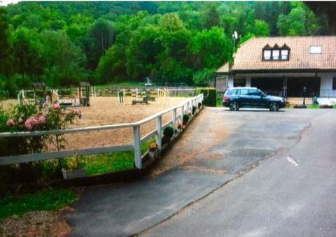 Ein Reitplatz mit Hindernissen und ein angrenzendes Gebäude sind sichtbar. Die Umgebung ist grün und von Bäumen umgeben.