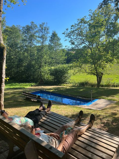 Zwei Personen liegen entspannt auf Liegen in der Natur. Im Hintergrund ist ein Swimmingpool und grüne Wiesen zu sehen.