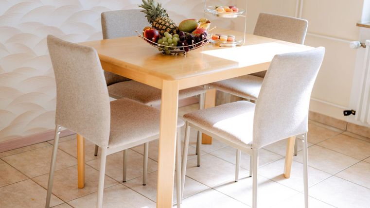 Een lichte eetruimte met een houten tafel en vier stoelachtige stoelen. Op de tafel staat een fruitmand met verschillende vruchten.