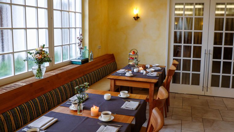 Ein gemütlicher Essbereich mit Holztischen und Stühlen. Die Fenster lassen viel Licht herein, und auf den Tischen stehen Blumen und Kerzen.