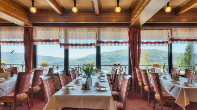 Ein eleganter Speisesaal mit Tischen, die für Gäste gedeckt sind. Durch große Fenster hat man einen Blick auf eine grüne Landschaft.