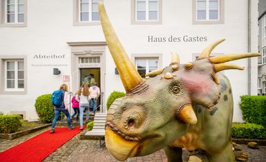 Ein lebendiges Dinosauriermodell steht vor dem Eingang zum "Haus des Gastes". Besucher gehen über einen roten Teppich hinein.