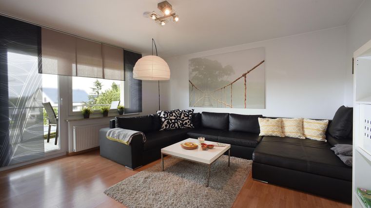 Ein modernes Wohnzimmer mit einem schwarzen Sofa, einem hellen Teppich und einem Tisch, der mit Snacks gedeckt ist. Große Fenster sorgen für viel natürliches Licht.