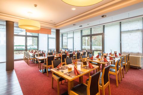 Ein modernes Restaurant mit Holzstühlen und Tischen. Die Einrichtung ist hell und einladend, mit großen Fenstern, durch die Tageslicht strömt.