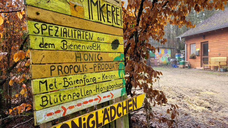 Ein bunter Holzschild mit Informationen über Imkereiprodukte steht vor einem kleinen Verkaufsladen. Die Umgebung ist von Bäumen und Herbstblättern geprägt.