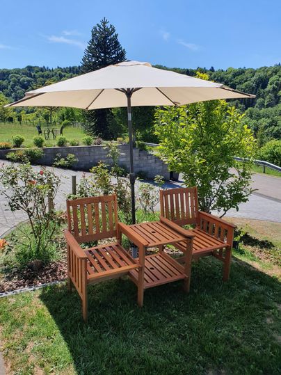 Zwei Holzstühle unter einem Sonnenschirm stehen in einem grünen Garten. Im Hintergrund sind Bäume und Sträucher zu sehen.