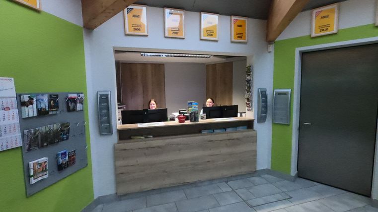 Ein Empfangsbereich mit zwei freundlichen Mitarbeiterinnen hinter einem modernen Empfangstresen. Die Wände sind in frischen Farben gestrichen, und es gibt Informationsmaterialien an der Seite.