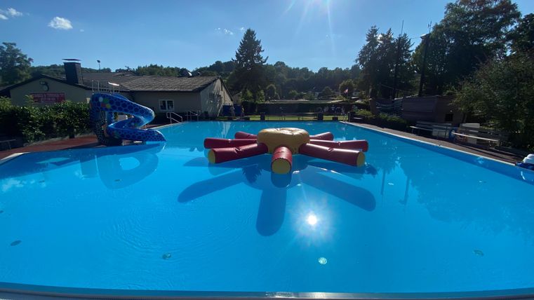 Ein großer Pool mit klarem blauen Wasser und einem bunten, aufblasbaren Spielzeug in der Mitte. Im Hintergrund sind Bäume und ein Gebäude zu sehen.