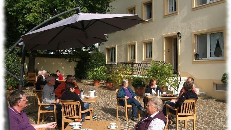 Eine Gruppe von Menschen sitzt gemütlich unter einem Sonnenschirm auf einer Terrasse. Es gibt Tische mit Kaffeetassen und eine ruhige, einladende Atmosphäre.