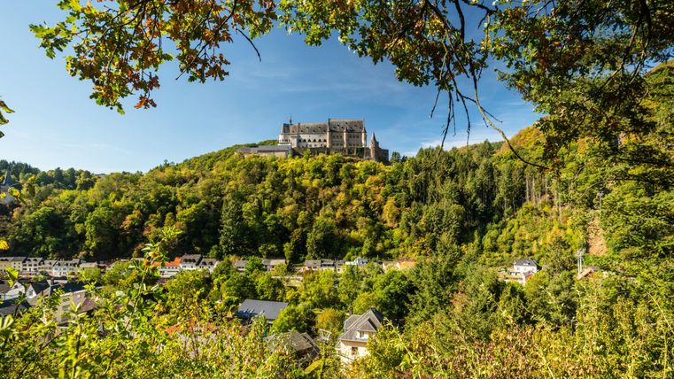 Eine beeindruckende Burg auf einem Hügel, umgeben von buntem Laub und Bäumen. Im Vordergrund sind kleine Häuser und eine grüne Landschaft zu sehen.
