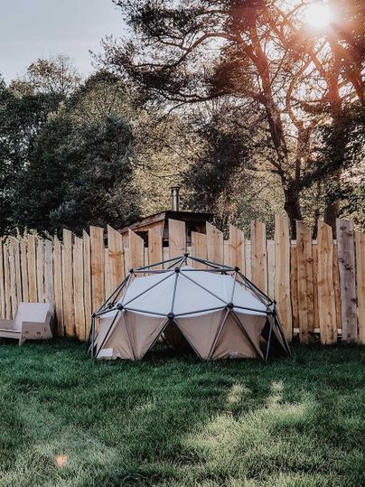 Ein modernes Geodätisches Zelt steht in einem Garten umgeben von einem Holzzaun. Im Hintergrund sind Bäume und ein sanfter Sonnenuntergang zu sehen.