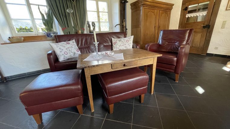 Ein gemütlicher Wohnbereich mit einem roten Ledersofa und zwei passenden Sesseln. Auf dem Holztisch liegt eine Tischdecke, und die Fenster lassen viel Licht herein.