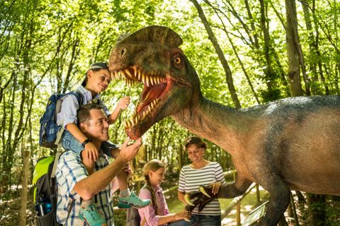 Eine Familie steht in einem Wald und interagiert mit einem Dinosaurier-Modell. Die Kinder sind begeistert, während die Erwachsenen lächeln und mit dem Dinosaurier spielen.