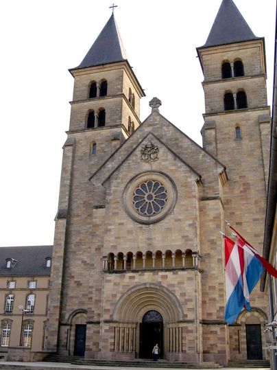Eine beeindruckende Kirche mit zwei Türmen und einer großen Rosette. Vor der Kirche wehen die Flaggen von Luxemburg.