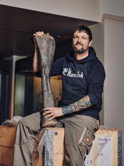 Een man zit op een houten kist en houdt een groot fossiel in zijn hand. Hij draagt een zwarte trui en heeft getatoeëerde armen.