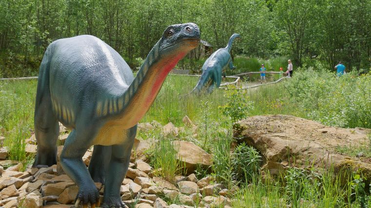 Zwei Dinosauriermodelle stehen in einem grünen Park. Die Landschaft ist mit Bäumen und Steinen geschmückt.