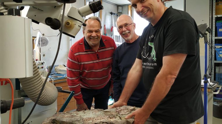 Trois hommes se tiennent dans un espace de travail et sourient. Ils tiennent un grand objet en pierre, tandis que des machines et des outils sont visibles à l'arrière-plan.