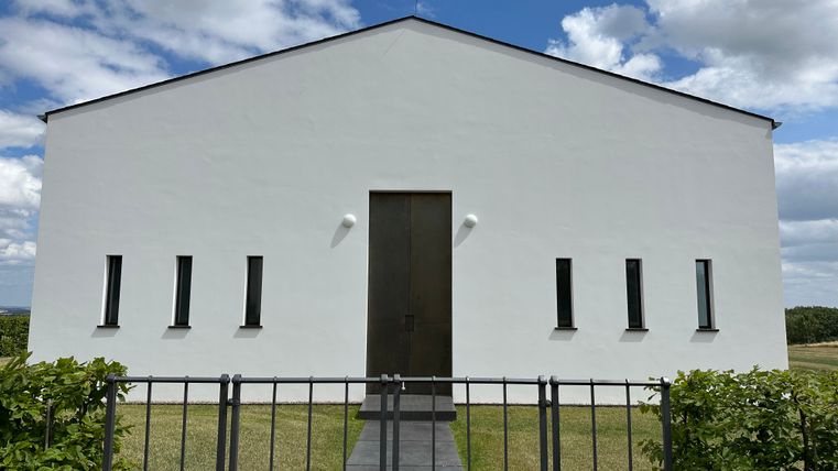 Modernes weißes Gebäude mit schmalen Fenstern und einem großen Holztor, umgeben von Rasen und einem Zaun, unter blauem Himmel mit Wolken.