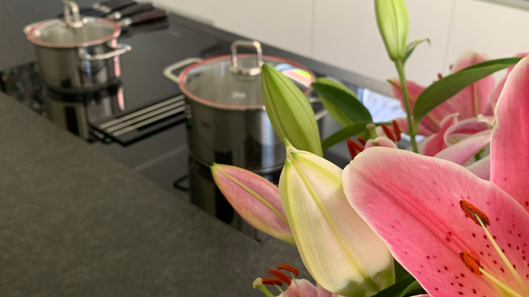 Eine moderne Küche mit einem schwarzen Kochfeld und zwei Töpfen. Im Vordergrund blühen rosa Lilien.