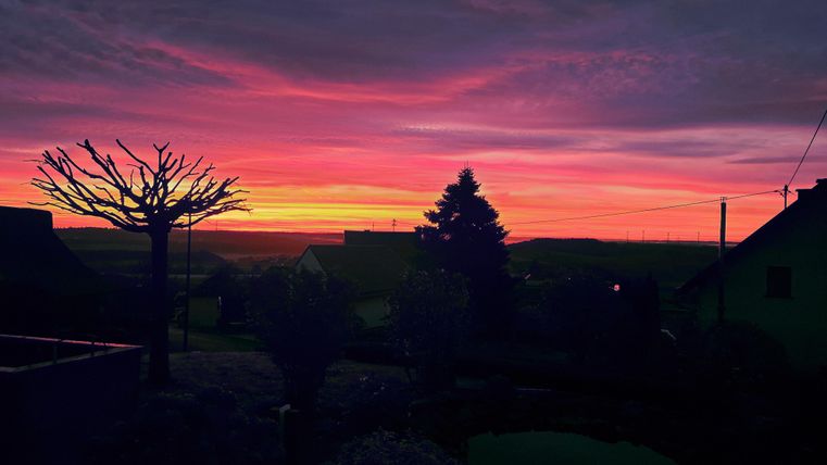 Ein wunderschöner Sonnenuntergang mit leuchtenden rosa und orangefarbenen Wolken. Silhouetten von Bäumen und Häusern verleihen der Szene eine ruhige Atmosphäre.