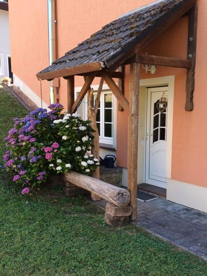 A charming entryway with a covered wooden porch. Colorful hydrangeas bloom next to the door, giving the house a welcoming charm.