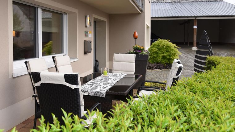 Ein gemütlicher Außenbereich mit einem Essplatz aus dunklem Rattan und einem Tisch. Grünes Licht und Pflanzen umgeben den Bereich, ideal für Mahlzeiten im Freien.