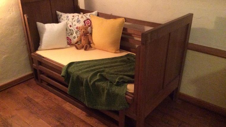 Ein gemütliches, helles Zimmer mit einem hölzernen Bett. Das Bett ist dekoriert mit bunten Kissen und einer grünen Decke.