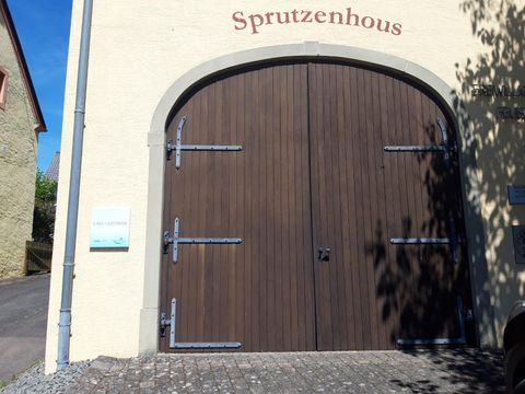 Eine große, geschlossene Holztür mit metalischen Beschlägen. Darüber steht das Wort "Sprutzenhaus".