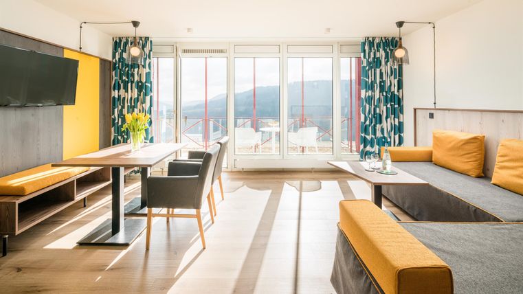 Ein modernes Wohnzimmer mit einem großen Fenster und Blick auf die Landschaft. Die Einrichtung ist hell und freundlich, mit einem Essbereich und einer gemütlichen Couch.